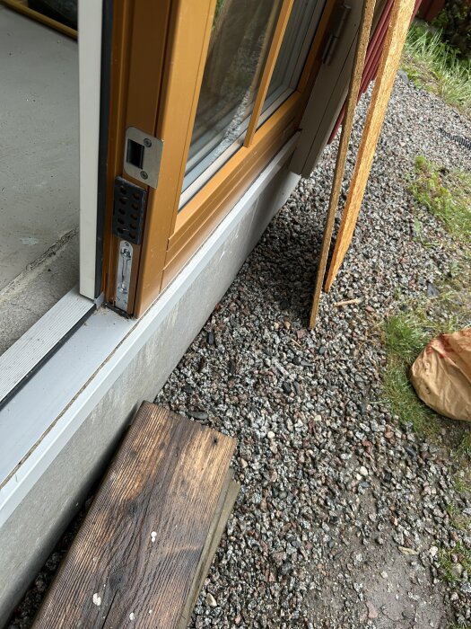 Öppen dörr med skadad dörrtröskel och plåt, blottande en glipa där vatten kan tränga in; grus och en träbräda synliga framför.