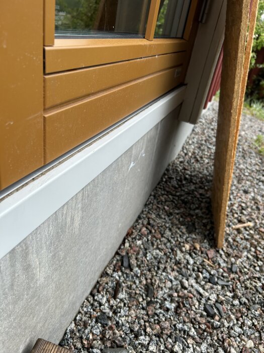 Dörrtröskel med plåt under, där plåten verkar ha fått en buckla och inte sluter tätt mot dörren, på en yttervägg med grus på marken.