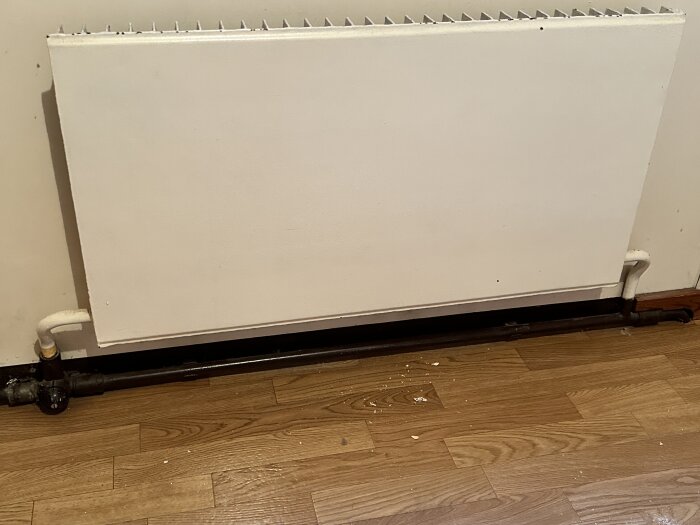 Radiator med anslutande rör och skarvar mot en vägg, placerad ovanför ett trägolv.