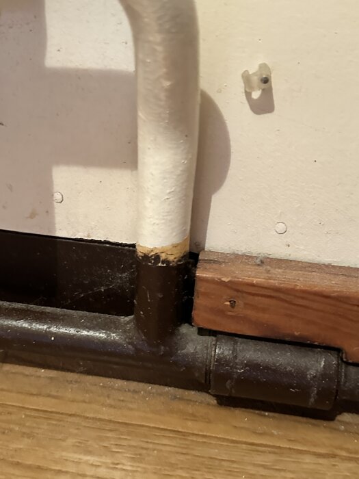 Rörledning med skarv och ett fäste mot en vägg med trälist.