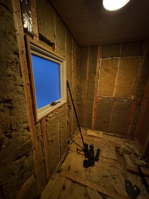 Renovering av badrum med exponerad isolering, verktyg på golvet och ett fönster. Träreglar och råspont syns på väggarna.