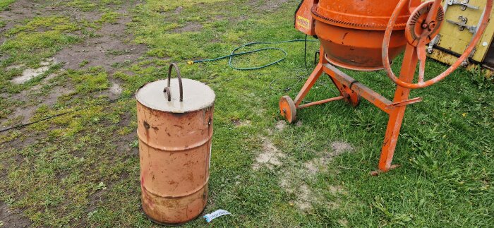 Rostig metallcylinder med betong i toppen och stålhandtag, står på gräs bredvid en cementblandare.