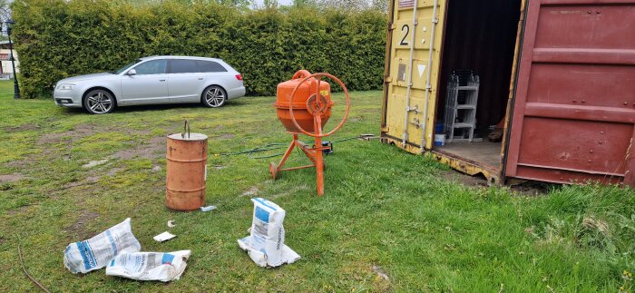 Cementblandare på gräs bredvid gul container, säckar med grovbetong och en metalltunna. Silverfärgad kombi i bakgrunden.