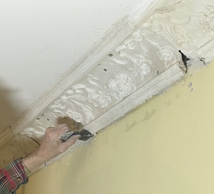 En hand använder ett stuckaturjärn för att applicera lim och spackel på en dekorativ taklist med blommönster.