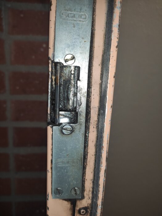 Elslutbleck installerat i dörrkarm, märkt med "Solid". Skruvar synliga, beläggning aningen sliten. Dörromgivning i flerbostadshus.