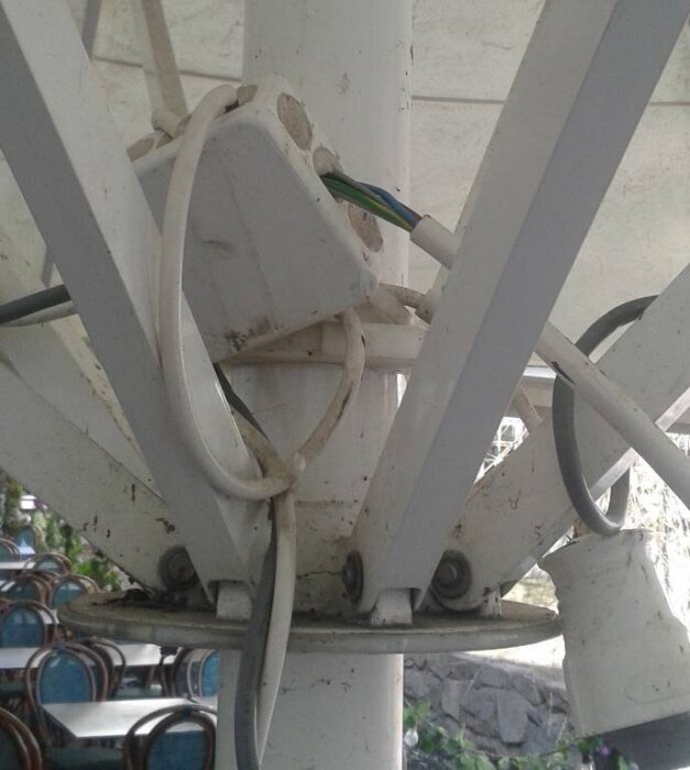 Detalj av parasollstolpe med kablar och elutrustning på en restaurangs uteservering, omgiven av stolar och bord.