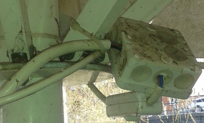 Elanslutning på parasollstolpe med flera sladdar och en kopplingsdosa, tecken på slitage och smuts.