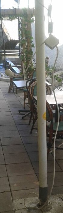 Parasollstolpe på restaurangs uteservering med kablar fästa, infravärmare hängande, stolar och bord synliga i bakgrunden, oordnade sladdar på marken.