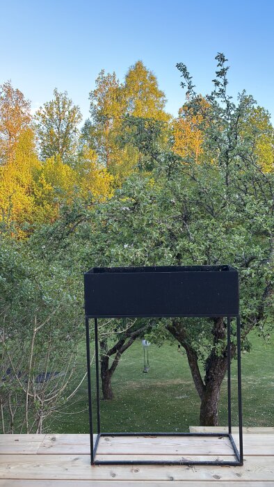 Svart blomlåda på metallstativ står på altan med grönskande träd och gräsmatta i bakgrunden.