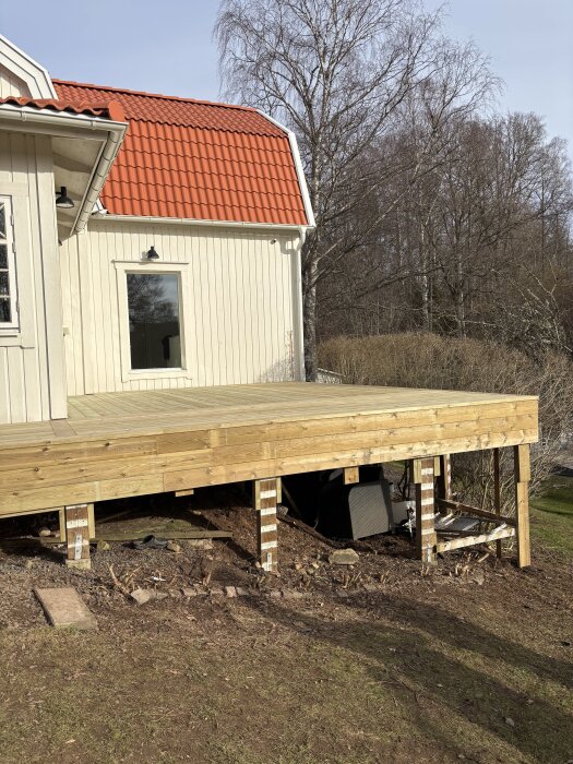 Träaltan under uppbyggnad fäst vid ett hus med rött tak, utan synligt staket, med en svart kruka placerad under.