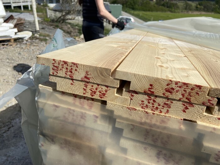 Staplad kärngran med röda markeringar, plastöverdragen, utomhusmiljö i bakgrunden, person ses i ena hörnet.