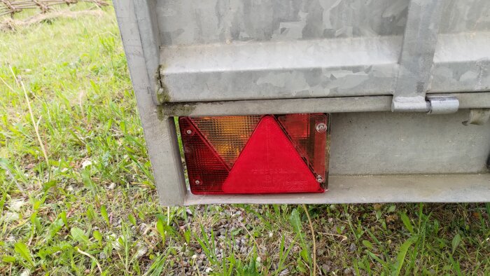 Bakljus på ett släp av märket Fogelsta, med röd triangelreflektor och orange blinkersglas, omgiven av gräs.