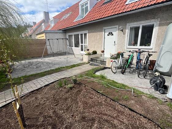 Småhus med röd takpanna, cyklar och betongplattor vid entrén, naken jord och planteringar. Bakgård med stensatt uteplats och lekställning syns.