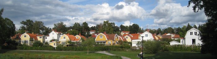 Gult och vitt hus från 2017, del av radhusområde med röda tak. Lummig omgivning med träd. Persona på bänk i förgrunden. Molnig himmel.