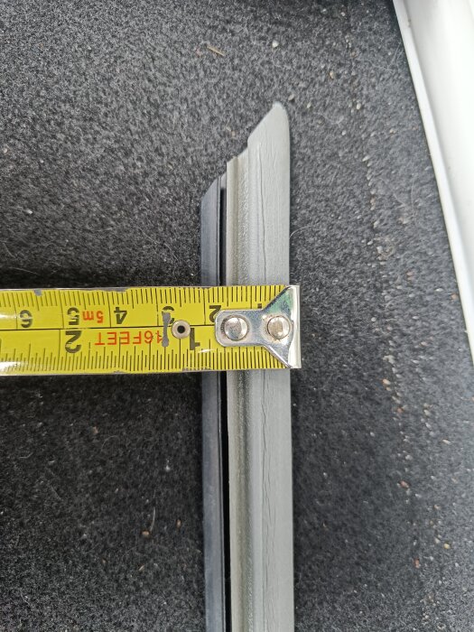 Mätning av grå fönstertätningslist med måttband; list placerad på mörk yta, listens längd är cirka 3,5 cm.
