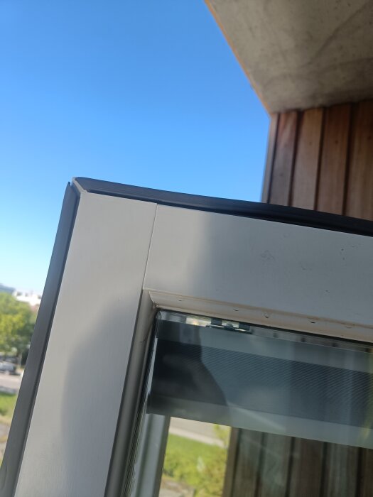 Närbild av hörnet på en balkongdörr som visar en tätninglist infälld i dörrens ram, utomhusmiljö med blå himmel i bakgrunden.