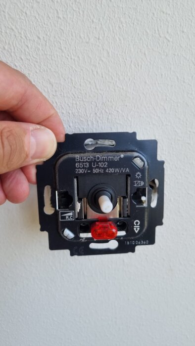 Busch-dimmer 6513 U-102 visas mot en vit vägg, hålls av en hand, märkningar och kopplingar synliga.