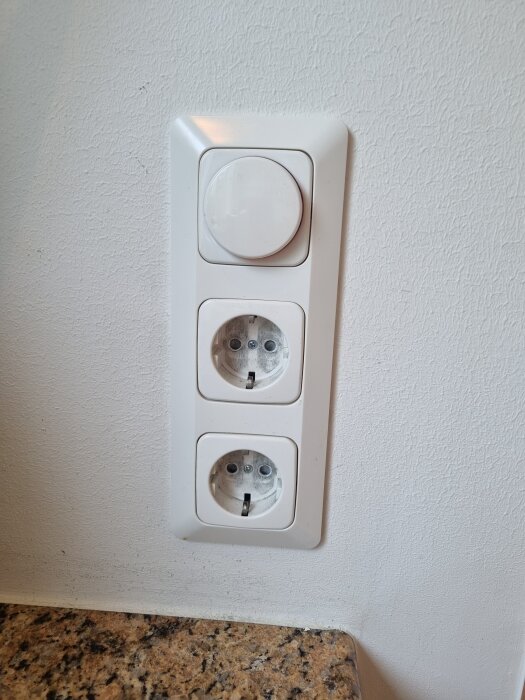 Vit väggpanel med en rund dimmer ovanför två eluttag på en vägg vid en köksbänk i sten.