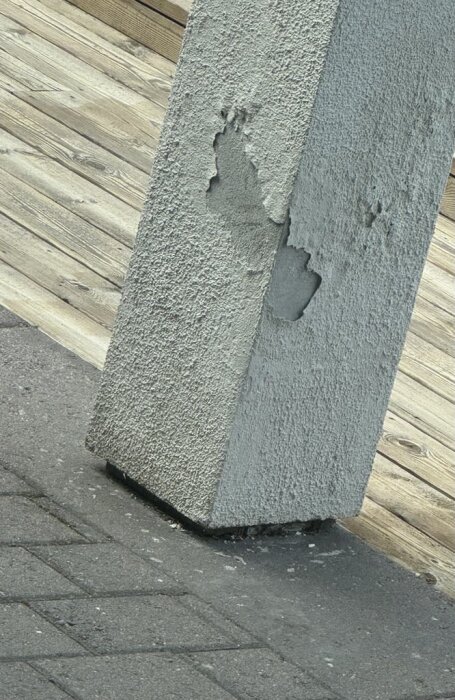Betongpelare med avflagnad puts, synlig på en trätrall och stengång, visar tecken på fuktskador och slitage.