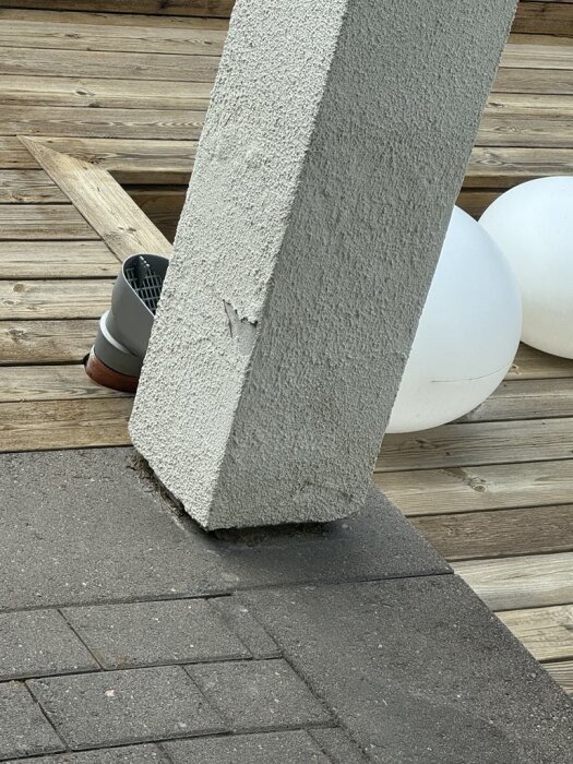 En putsad pelare med märkbar skada vid basen, står på ett trädäck. Det finns också synliga vita klotformade lampor i bakgrunden.