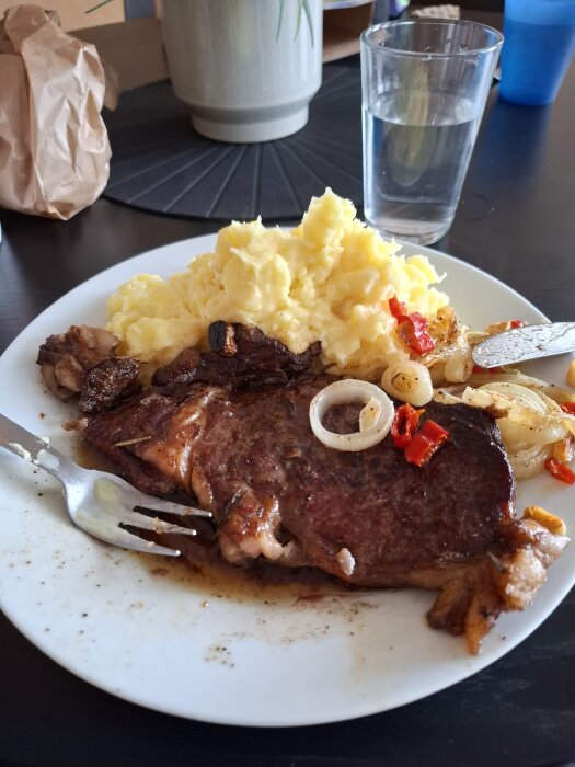 Tallrik med potatismos, en skiva entrecote och lök, serverad med ett glas vatten i bakgrunden.