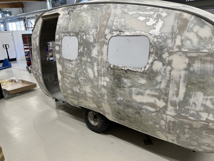 Husvagn under renovering i verkstad, med nya fönsterramar monterade. Golv och verktygsställ synliga i bakgrunden.