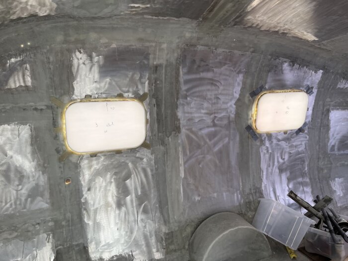 Två nyinstallerade, ovala fönster i en husvagnsvägg, fästa med limning och fastplastning, omgivna av en slät, metallisk yta och verktyg på golvet.