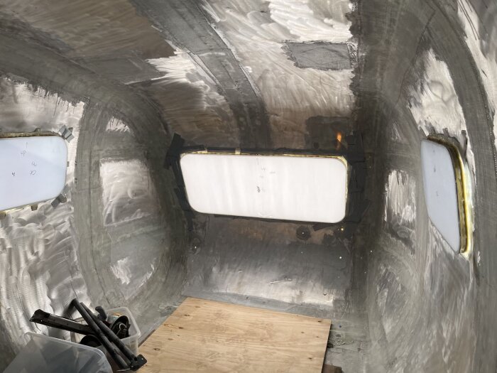 Insidan av en husvagn med fönster monterade på väggarna, delvis täckta med material runt omkring; golvet är av plywood.