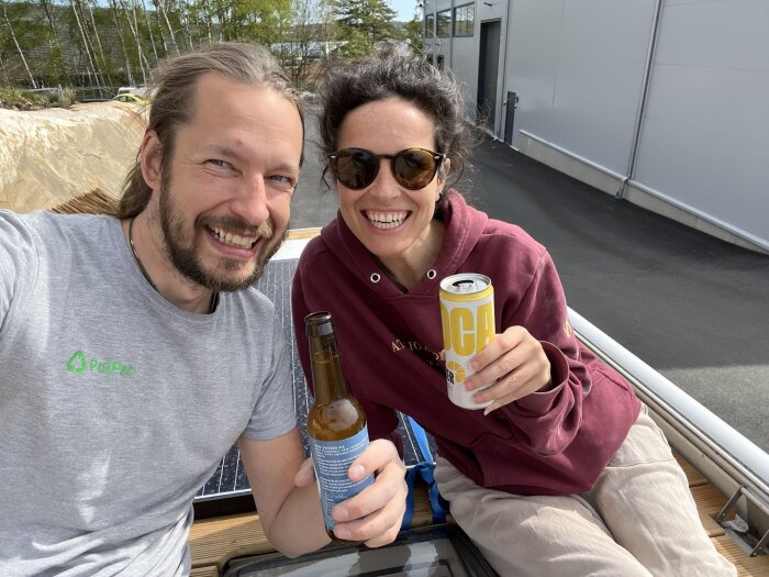 Personer skålar med dryck på ett tak eller plattform, soligt väder. En man i grå tröja och en kvinna i solglasögon och vinröd tröja ler mot kameran.