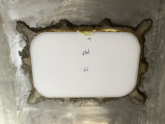 Ett insatt fönster i en husvagnsvägg, omgiven av applicerat lim och glasfiber för fastsättning, med handskriven text på skyddsplasten.