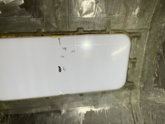 Rektangulärt fönster monterat med glasfiber och lim, med text som markerar riktningen. Bakgrund av metallplåt, del av husvagnsrenovering.