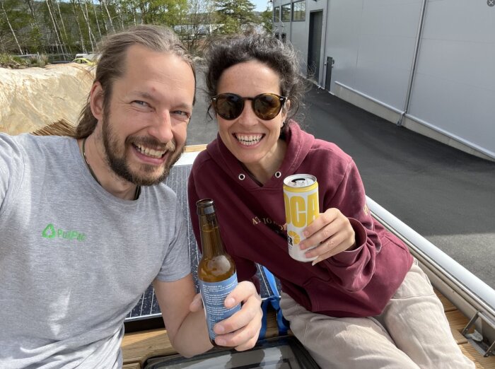 Två personer ler och håller drycker medan de sitter utomhus på ett fordon, omgivna av byggnader och grönska i bakgrunden.