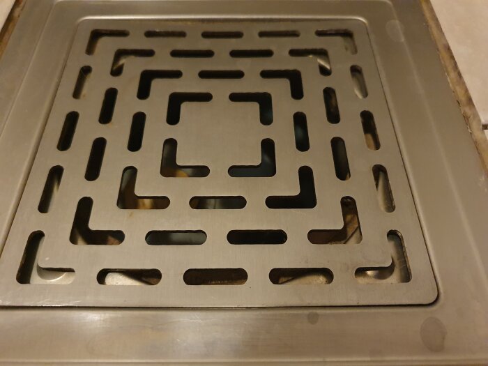 Brunnslock i dusch av metall med rutmönster, del av golv syns runt om; personen undrar hur man öppnar locket för rensning.