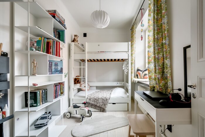 Barnrum med våningssäng, bokhylla fylld med böcker och leksaker, skrivbord med laptop och hörlurar. Färgglada gardiner vid fönstret.
