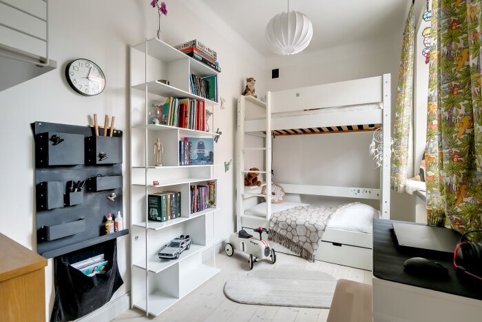 Barnrum med våningssäng, hylla med böcker och leksaker, samt en leksaksbil på golvet. Färgglada gardiner och en skrivbordsstation syns också.