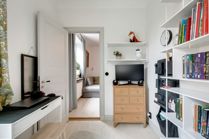 Barnrum med skrivbord, bokhylla och byrå, dörr öppen mot ett angränsande rum med soffa.