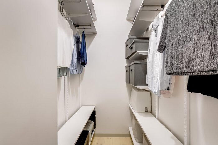 Walk-in closet med klädhängare, skjortor och klädlådor på hyllor. Organiserat och tömt för fotografering.