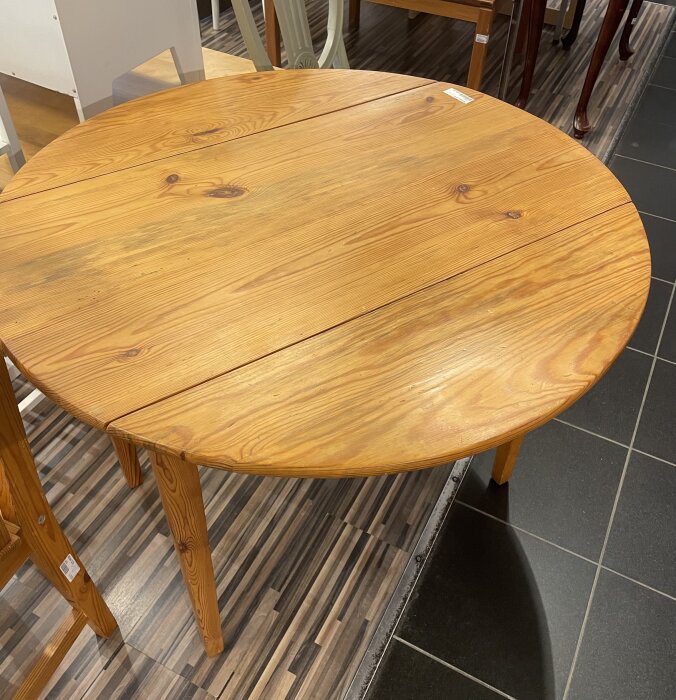 Runt matbord i trä med lackad yta, står på ett golv av mörka och ljusa klinkerplattor. Bordet har fyra ben och en synlig skarv på skivan.