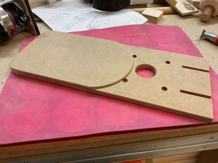 Träverktyg med slitsar och ett cirkulärt hål, gjord av MDF, placerad på en arbetsbänk med ritningar och verktyg i bakgrunden.