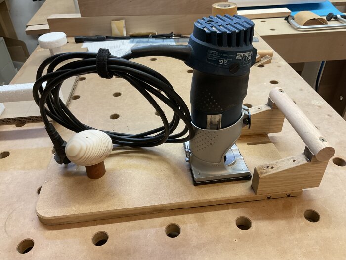 En handhållen fräs monterad på en träbas med ett handtag och en knopp, placerad på ett arbetsbord med hål.