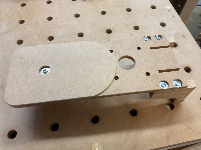Ett hemmabyggt verktyg i MDF med hål och slitsar, lagt på en arbetsbänk med hål, del av ett snickeriprojekt.