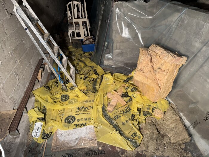 Oordnad källare med stege, staplade stolar, gul plast och isoleringsmaterial på golvet.