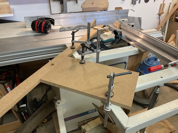 En arbetsbänk med fastklämd MDF-skiva och hörselskydd liggande brevid, omgiven av träbearbetningsverktyg i en verkstad.