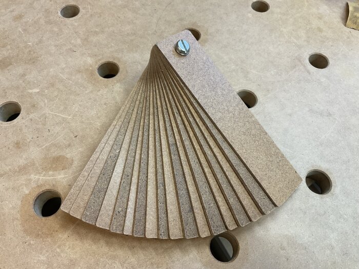 Fiberskivremsor med skruv, används som justerbart mellanlägg i träbearbetning. Ligger på en arbetsbänk med hål.