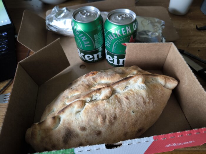Calzone i en kartong bredvid två burkar öl och en inlindad kebabrulle på ett bord.