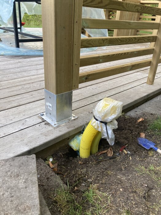 Gula slangar vid en trästolpe på en altan, delvis täckta med plast, sticker upp ur marken bredvid jord och verktyg.