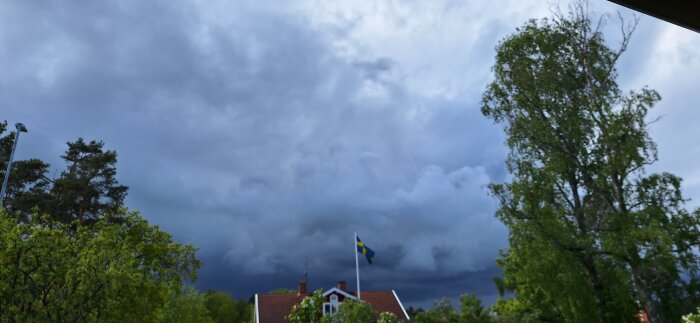 Hus med rött tak under mörka moln, svenska flaggan vajar, omgiven av gröna träd.