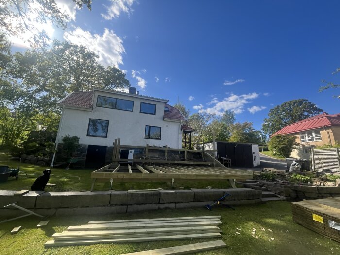 Tvåvåningsaltan under konstruktion framför ett vitmålat hus, med brädor och verktyg utspridda på gräsmattan.