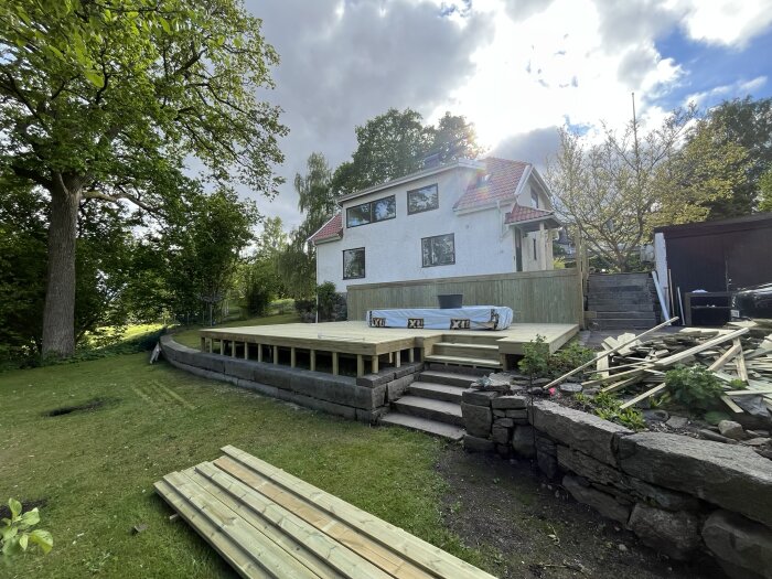 Två nivåers altan under konstruktion med grovträsteg, omgiven av grönska och stenar. Hus i bakgrunden till vänster.