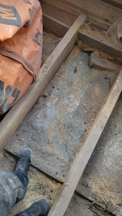 Spricka i betonggolv under renovering av 30-talshus, omgiven av träbjälkar och byggdamm. En person står med arbetsskor bredvid sprickan.